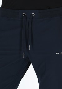 Pantaloni jogger blu navy con vita elastica, dettagli con coulisse e punte metalliche argento. Presentano un piccolo logo bianco sul lato.