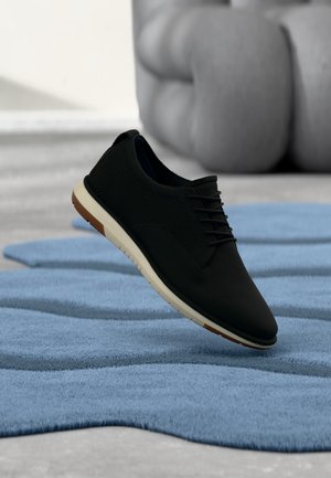 Scarpa casual nera con lacci e suola bianca che fluttua sopra un tappeto blu strutturato in un ambiente interno minimalista.