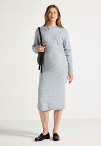 Gravid kvinna i en grå stickad tröja och klänning, som håller en svart väska, leende mot en neutral bakgrund.