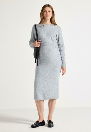 Gravid kvinna i en grå stickad tröja och klänning, som håller en svart väska, leende mot en neutral bakgrund.
