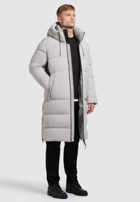 Grau gepolsterter Mantel mit Kapuze, Reißverschluss, Seitentaschen und texturiertem Finish, kombiniert mit schwarzen Hosen und Schuhen.