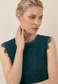 Joven mujer con cabello rubio, vestida con una blusa sin mangas de encaje color verde azulado, tocándose el cuello y mirando pensativamente hacia un lado sobre un fondo beige.