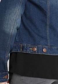 Denim takki, joka on valmistettu tummansinisestä kankaasta, jossa on haalistunut rakenne, ruskeat metallinapit ja musta alaosa. Yksinkertainen tyyli, jossa on näkyvät saumat.