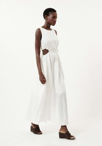 Robe blanche, longueur cheville, avec un corsage ajusté, découpes latérales et accents à cordon. Le modèle porte des sandales compensées en marron. Texture lisse.
