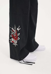 Zwarte cargo broek met een opvallend rood, wit en grijs geborduurd ontwerp op het linkerpijpje; gecombineerd met witte sneakers.