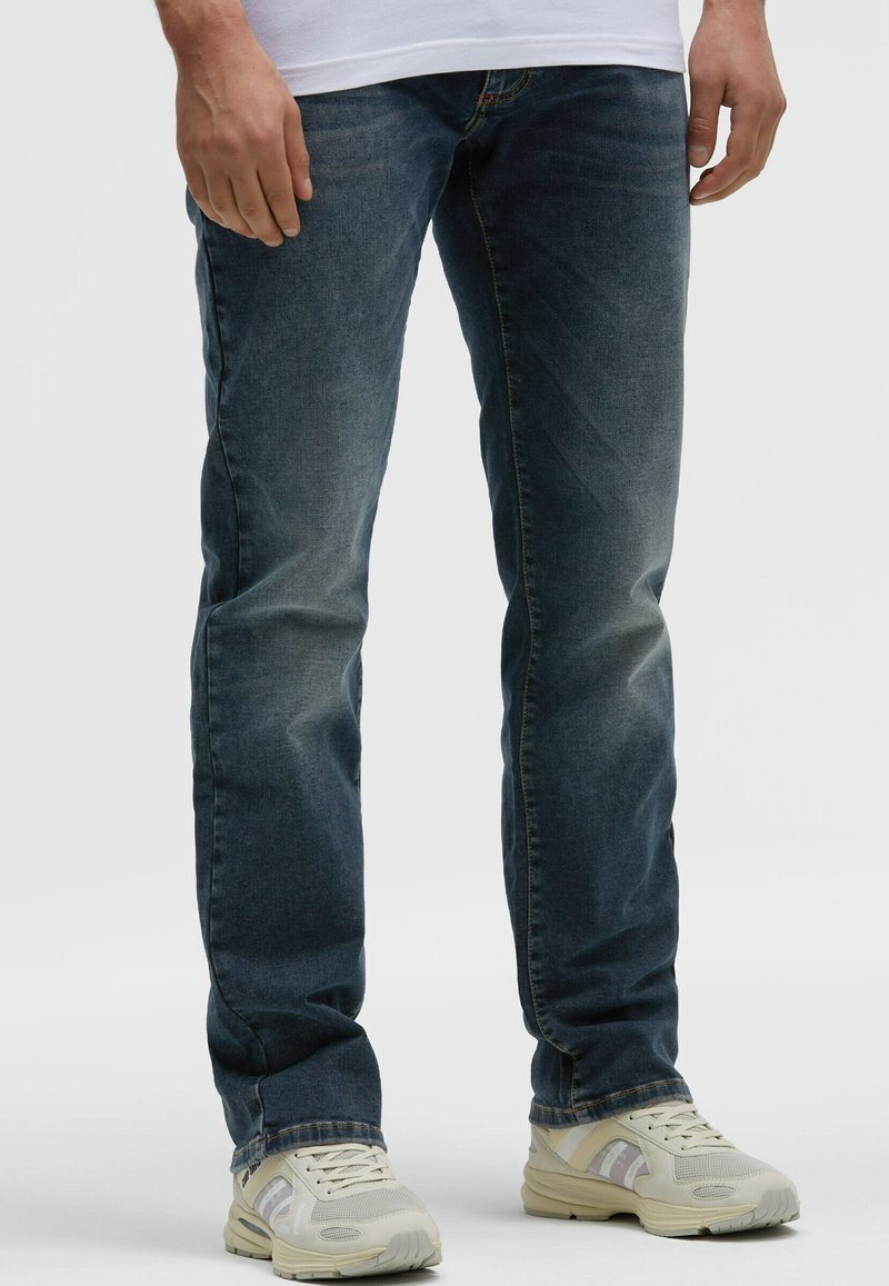 Mann trägt ausgewaschene blaue Jeans und weiße Turnschuhe, steht vor einem schlichten hellen Hintergrund.