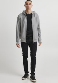 Sweat à capuche gris zippé avec cordon de serrage, associé à un t-shirt noir, un pantalon slim foncé et des baskets noires. Design simple et décontracté.