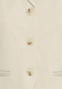 Tissu de couleur crème avec trois boutons brillants, ronds, à quatre trous, alignés verticalement sur un vêtement avec deux poches horizontales.
