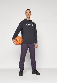 Sweatshirt noir à capuche avec logo, assorti à un pantalon violet et des baskets noires. La personne tient un ballon de basket orange.