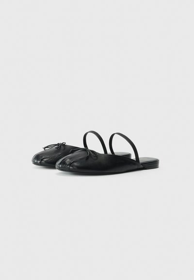 Black leather slide sandals with a rounded toe design, flexible straps, and a small decorative bow on the front. Flat sole.