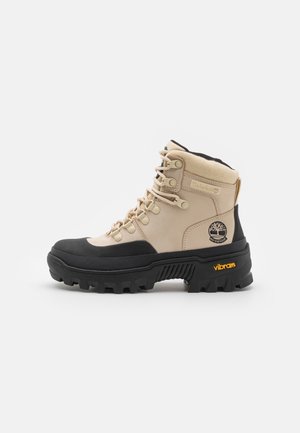 Botte de randonnée imperméable Timberland beige et noire avec une semelle Vibram épaisse et un laçage à l'avant, vue de côté sur fond blanc.