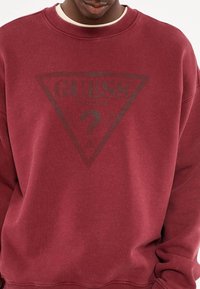 Bordeaux sweatshirt met ronde hals, voorzien van een donkere driehoek en de tekst "GUESS" in het midden. Zachte textuur en een ontspannen pasvorm.