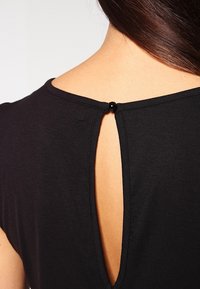 Top nero con dettaglio a chiave sul retro, fissato con un piccolo bottone nero, realizzato in tessuto morbido e liscio con una lieve texture.