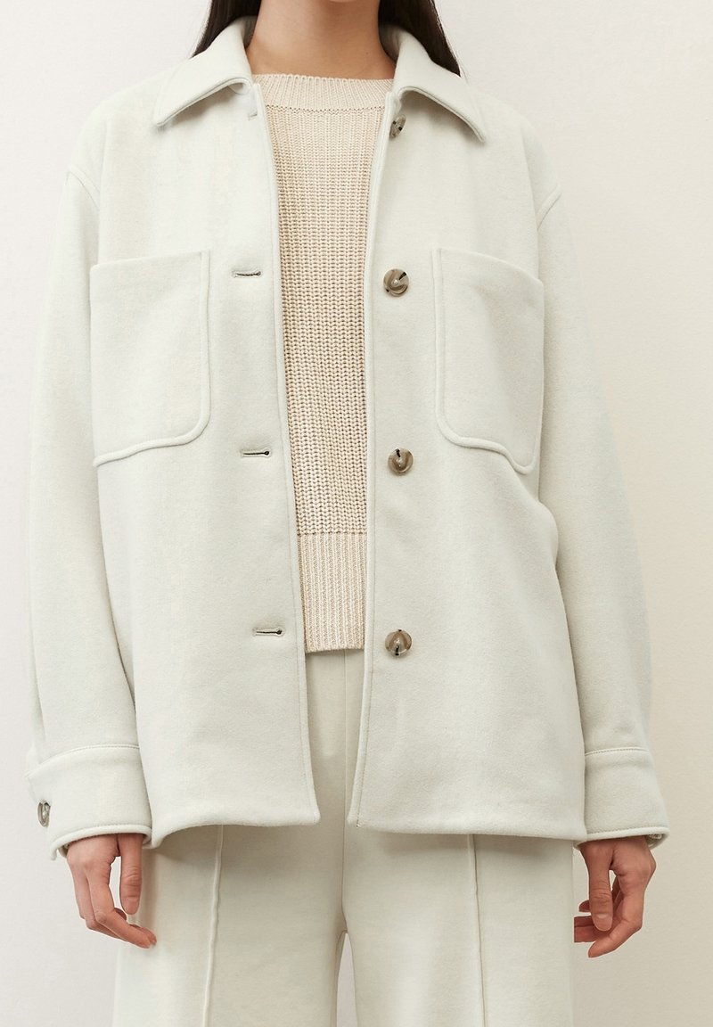 Femme portant une veste en laine écrue boutonnée par-dessus un pull en tricot beige et un pantalon écru assorti, debout devant un fond uni.