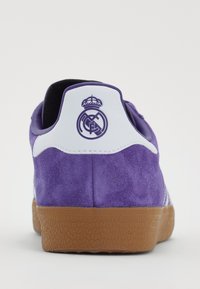 Sapatilha de camurça roxa com sola de borracha branca e de cor de borracha. Apresenta um logo no calcanhar com um detalhe de coroa, material texturizado e acabamentos cosidos.