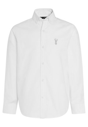 Chemise blanche à manches longues, boutonnée, avec col et petit logo de tête de cerf brodé sur la poitrine gauche. La chemise est soigneusement repassée.