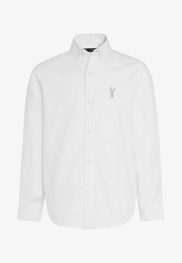 Chemise blanche à manches longues, boutonnée, avec col et petit logo de tête de cerf brodé sur la poitrine gauche. La chemise est soigneusement repassée.