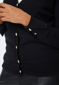 Cardigan nero realizzato in tessuto morbido con maniche lunghe. Presenta dettagli con bottoni dorati e polsini a coste, che aggiungono texture e particolari.