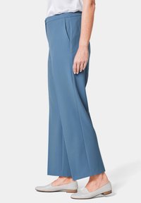 Hoch geschnittene, weite Hose aus weichem blauen Stoff, mit vorderen Falten und seitlichen Taschen. Kombiniert mit hellgrauen Ballerinas.