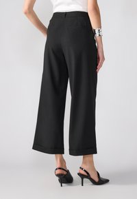 Svarte vide bukser med glatt tekstur, avkorte hem og to baklommer, parret med svarte slingback-heler.