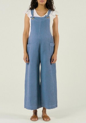 Femme portant une salopette large à jambes larges texturée bleue avec de grandes poches avant, un chemisier blanc à manches courtes et des sandales marron à enfiler.