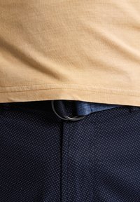 Tricou din bumbac galben auriu, asortat cu pantaloni din țesătură bleu închis, cu un model punctat. O inel metalic negru este așezat pe o curea albastră.