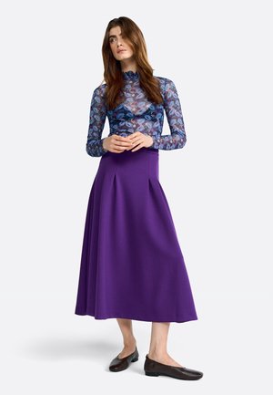 Lunga gonna plissettata viola abbinata a una blusa azzurra trasparente con motivo floreale. La modella indossa scarpe basse scure. Sfondo semplice.