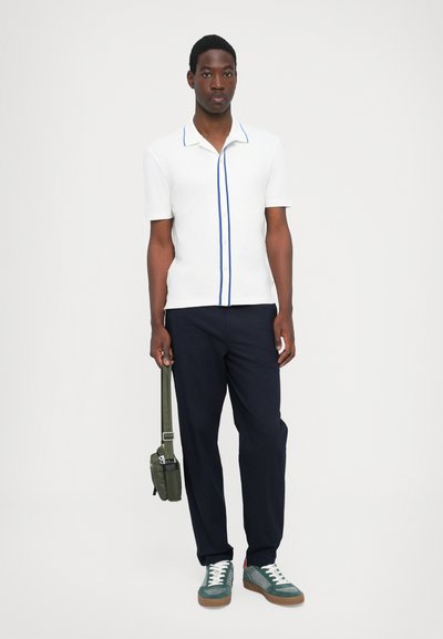 Cămașă albă cu mânecă scurtă și detalii albastre, pantaloni negri, adidași verzi, purtând o geantă de umăr verde. Design simplu și modern.