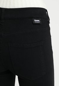 Svarta denimjeans med en bakficka som har en vävd etikett. Tyget ser slätt ut och sömmen är synlig längs med sömmarna.