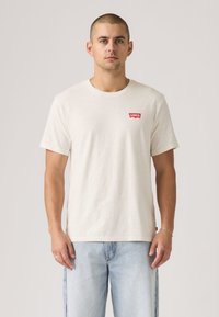 T-shirt en coton crème avec un patch logo Levi's rouge sur la poitrine ; accompagné d'un jean en denim bleu clair. Design simple à manches courtes.