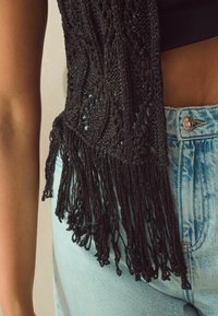 Gilet en tricot noir à détails en franges, présentant un motif à mailles larges, associé à un jean en denim clair taille haute.
