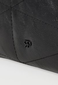 Superficie in pelle nera con cuciture diagonali e un piccolo logo rialzato che presenta lettere stilizzate "RR" vicino al bordo inferiore.