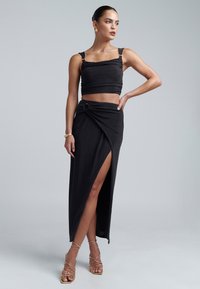 Crni dvodijelni outfit s prilagođenim topom s debelim trakama i suknjom visokog proreza, naglašenom detaljem u obliku uvijenog čvora i sjajnom tkaninom.