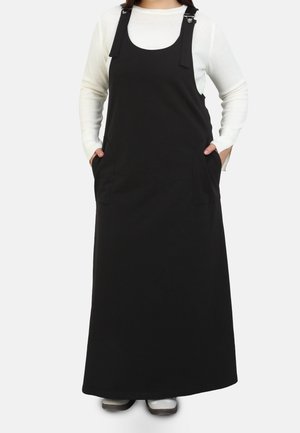 Person wearing a long black sleeveless dress with front pockets over a white long-sleeve top, standing with hands in pockets.