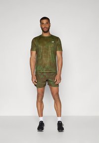 Conjunto desportivo com padrão camuflado verde, incluindo uma t-shirt justa e calções, feitos de tecido que afasta a humidade. Ténis pretos; meias brancas.