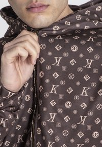 Sudadera marrón con cremallera y logotipos y patrones en beige claro, con una textura suave y capucha con cordón. Detalle de cremallera visible en la parte delantera.