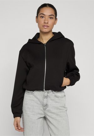 Noor tume juustega inimene, kes kannab musta lukuga kapuutsi ja heledaid halli teksapükse, seisab ühe käega taskus.