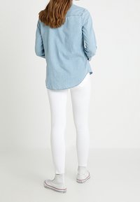 Camicia in denim azzurro chiaro con maniche lunghe, orlo curvato e dettaglio dei bottoni; abbinata a pantaloni bianchi aderenti e sneaker grigie.