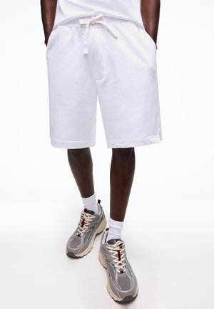 Mężczyzna w białych spodenkach ze sznurkiem, białych skarpetkach i szarych sportowych butach, stoi z rękami w kieszeniach.