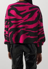 Donna che indossa un maglione astratto largo nero e rosa brillante e pantaloni neri lucidi con texture, mostrata di spalle.