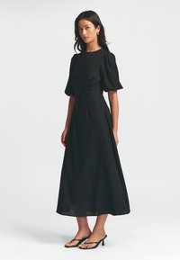 Schwarzes Midi-Kleid mit taillierter Passform, Puffärmeln und glattem Stoff. Kombiniert mit schwarzen Sandalen mit dünnen Riemen und Absatz.