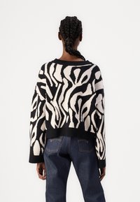 Svart og kremfarget genser med abstrakt mønster, løs passform, ribbede ermer og hem. Båret med mørkeblå jeans, sett bakfra.