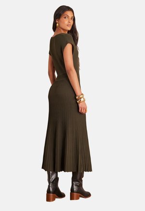 Robe en maille côtelée vert olive avec manches courtes et jupe évasée plissée. Associée à des bottes noires montantes et des accessoires dorés.