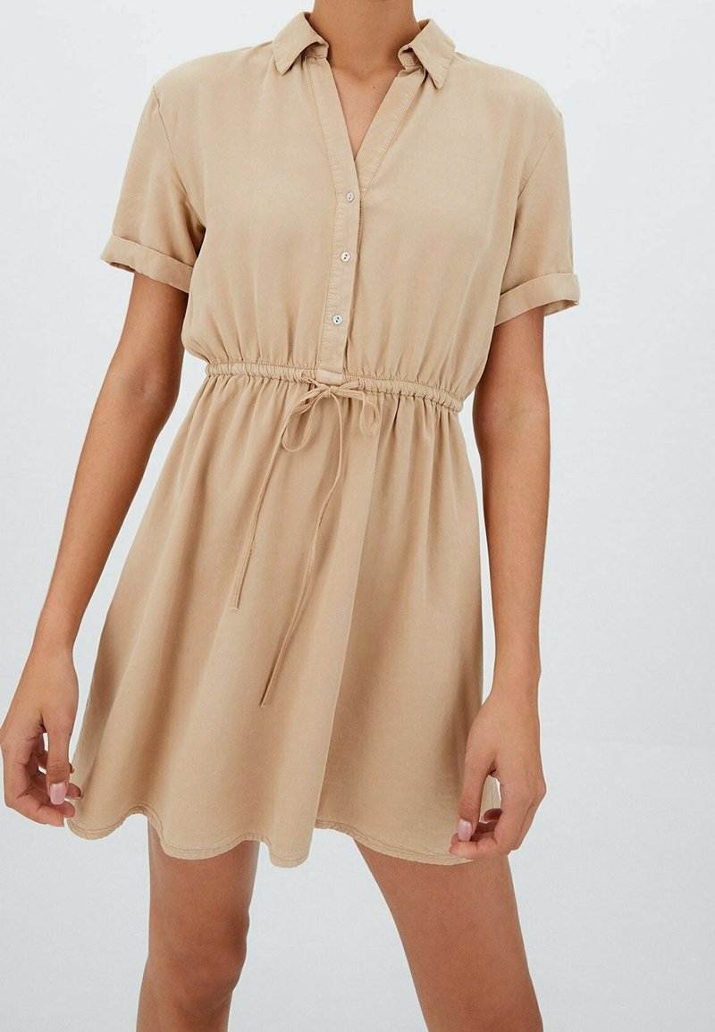 Sukienka w stylu koszulowym z krótkim rękawem, wykonana z jasnego beżowego materiału. Posiada guziki z przodu oraz sznurek w talii, z falbanistym dołem.