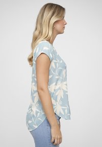 Blouse bleu à fleurs avec manches courtes, motif botanique blanc, encolure arrondie et ourlet asymétrique. Portée avec un jean bleu clair.