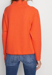 Jersey texturizado naranja con cuello alto y mangas raglán, combinado con vaqueros azules. La tela presenta un patrón tejido.