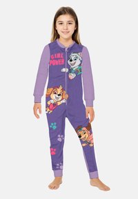 Ragazzina che indossa un pigiama intero viola con cani cartoon, impronte di zampe e il testo "GIRL POWER" sulla parte anteriore, in piedi a piedi nudi.