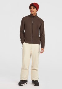 Brun fleecejakke med høj krave og frontlynlås, kombineret med beige cargo-bukser og sorte snesko. Rød strikhue fuldender looket.