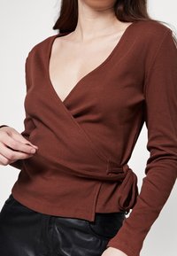 Mujer vestida con una blusa marrón de canalé anudada a la cintura, combinada con pantalones negros, sobre un fondo blanco liso.