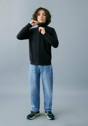 Veste polaire noire à fermeture éclair, jean bleu clair et baskets foncées. Cheveux bouclés, pose debout, fond gris uni.